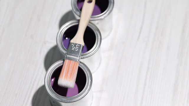 Cans with paint in shades of purple and brush on white wooden table, above view. Camera moving in