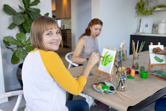 Two women smiling and painting on small tabletop easels at bright studio table with paintbrushes