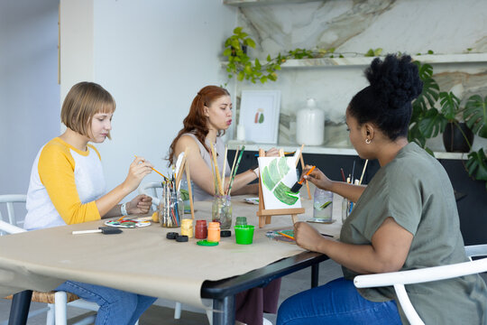 Three female friends painting at studio around papered table with easel, paint jars, smartphone