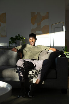 African American man sitting on gray sofa at home, wearing olive tee, plants casting shadows
