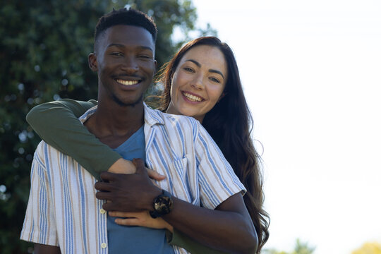 Diverse couple embracing, smiling in park, man in striped shirt and watch, woman in green top