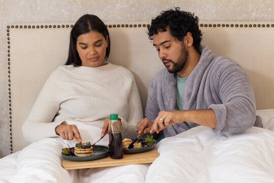 African American couple wearing sweater, robe sitting in bed sharing pancakes on tray with syrup