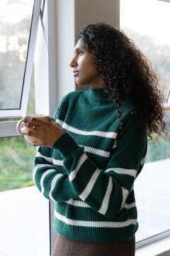 African American woman holding mug, wearing striped sweater by casement window showing gold ring