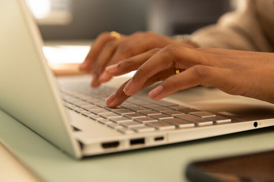 African American woman typing on laptop keyboard at desk, beige long-sleeves and gold ring visible