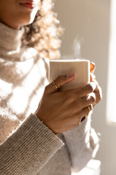 African woman holding steaming ceramic mug with gold rings, in ribbed turtleneck by window light