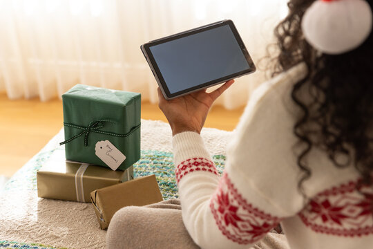 African woman wearing Nordic sweater using tablet on sofa at sunlit window beside gifts, copy space