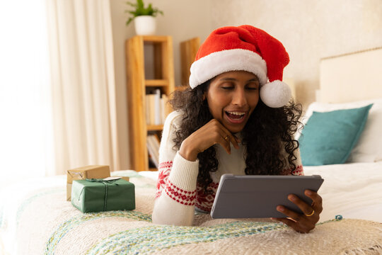 African American woman wearing Santa hat, lying on bed, holding tablet near wrapped gifts
