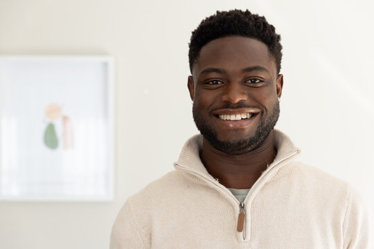 African man smiling while wearing beige zip-up pullover, stud earring at home beside artwork left