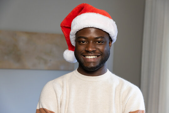 African man standing and smiling facing camera at home wearing Santa hat and cream knit sweater