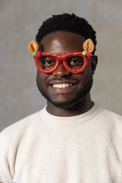 African adult male wearing cream sweater and red glitter glasses with pom-poms, smiling at backdrop