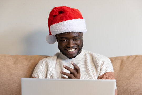 Adult African male wearing Santa hat, cream sweater, sitting on beige couch, waving to laptop