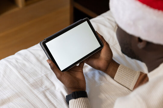 Adult African man wearing Santa hat, sweater and watch holding tablet sitting on bed, copy space