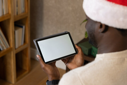 African American man wearing Santa hat holding tablet and showing blank screen in living room