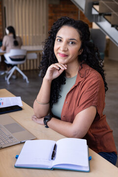 Female coworkers, woman wearing rust top working at wood desk in open office with laptop, notebook