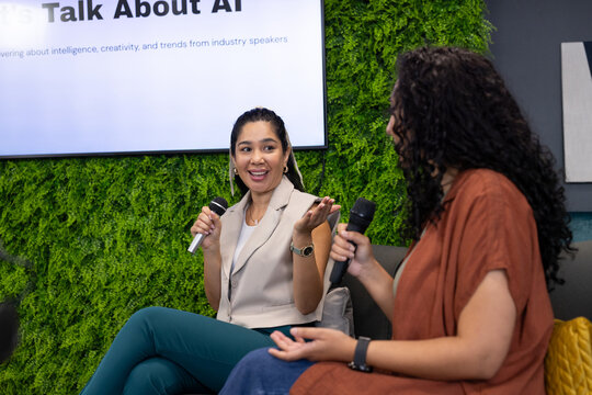 Diverse female panel wearing sleeveless blazer speaking on stage with microphones, plant wall