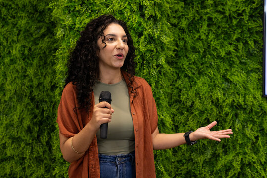 African American woman speaking into mic, gesturing by plant wall, showing watch, bangle, monitor