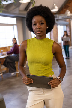 African American woman standing in coworking space holding tablet wearing green top and light pants