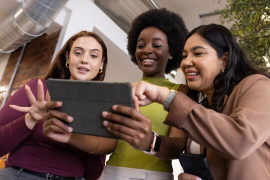Diverse female coworkers huddling, reviewing tablet case and phone in business casual office