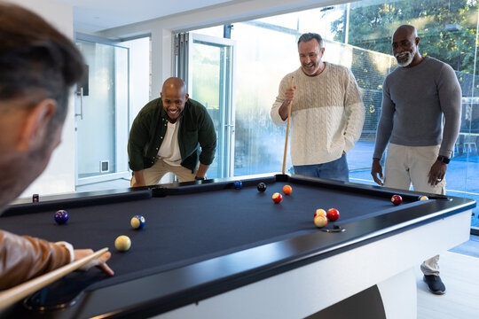Diverse male friends playing pool around black-felt table in modern recreation room with cues