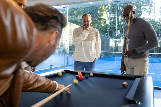 Diverse male friends leaning over black felt pool table in game lounge, brown jacket holding cues