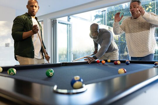 Diverse male friends lining up cue shot and reacting in home game room with navy-blue felt