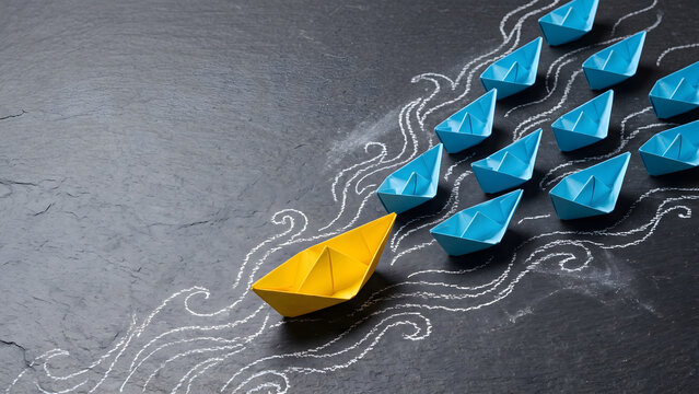 A bright yellow paper boat courageously leads a procession of blue boats, symbolizing innovative leadership and setting a new course for success and progress