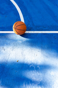 Orange basketball resting on blue concrete court at white line intersection, casting long shadow