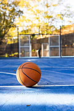 Orange basketball is sitting on blue outdoor court near white boundary line, casting long shadow
