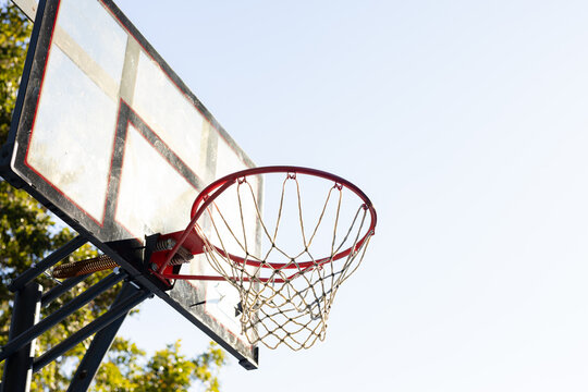 Freestanding basketball hoop is standing near trees under pale blue sky, showing frayed net
