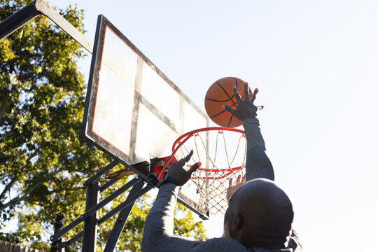 Mature African man in gray top reaching up at outdoor hoop holding orange basketball above rim