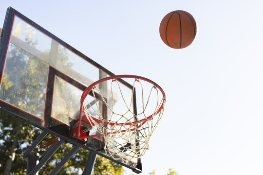 Basketball hoop and clear backboard are shining in sunny yard, orange ball hovering above rim