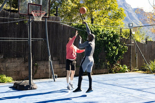 Two males jumping, shooting basketball in grey top, red sweatshirt, contesting hoop on blue court
