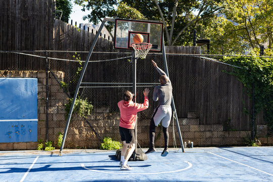 Diverse male friends playing basketball on blue backyard court, jumping toward hoop, bottles nearby
