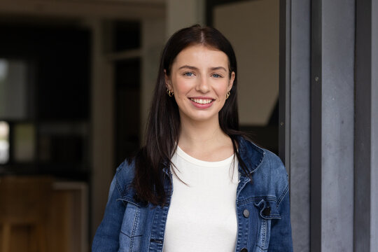 Woman standing smiling at home near doorway wearing denim jacket and gold hoop earrings