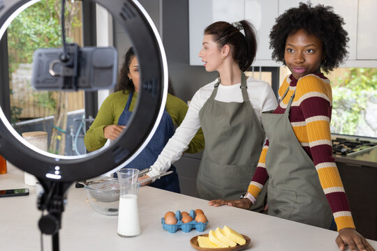 Diverse female friends in aprons making batter in bowl at kitchen counter filming with ring light