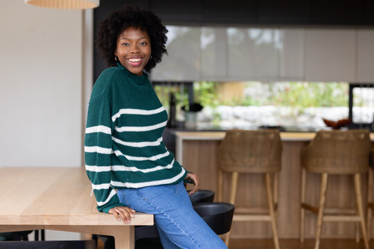 African American woman sitting on wooden dining table in kitchen smiling, wearing striped sweater
