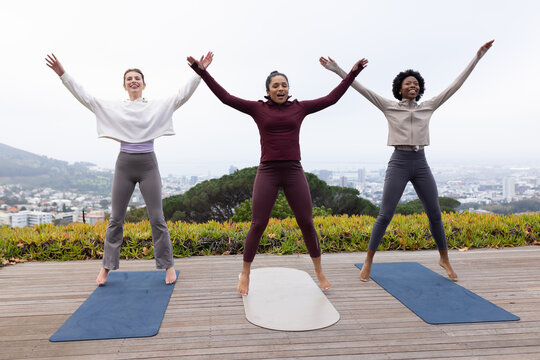 Diverse female friends in leggings, jackets performing jumping jacks on deck with three mats