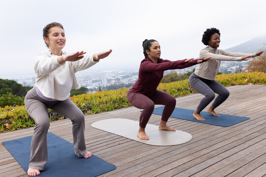 Diverse female friends in activewear holding low squat on three yoga mats on deck overlooking city