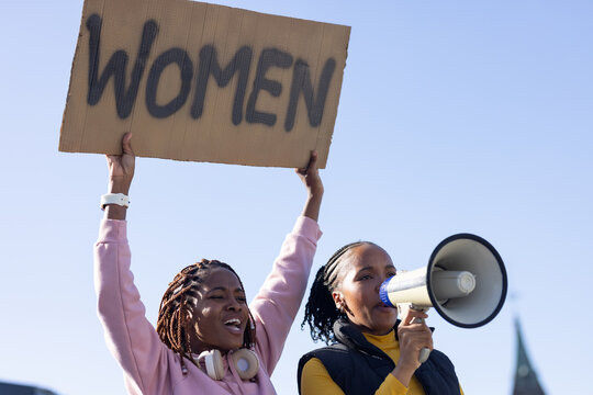 Two African women raising cardboard sign reading WOMEN and shouting into megaphone under blue sky