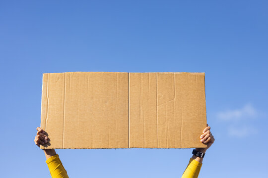 African American woman holding cardboard sign over head under blue sky, yellow sleeves, dark watch