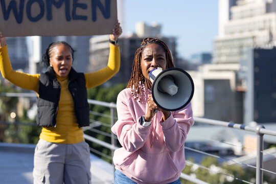 African women wearing pink hoodie leading rooftop protest, shouting into bullhorn with WOMEN sign