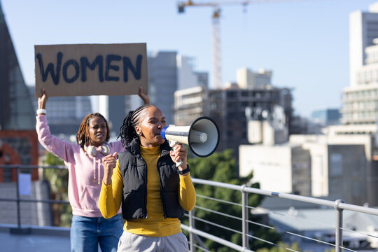 African women chanting with megaphone on rooftop by metal railing, in yellow top holding WOMEN sign
