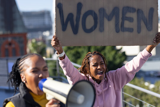 African women chanting, holding WOMEN sign on rooftop, pink hoodie and yellow top using megaphone