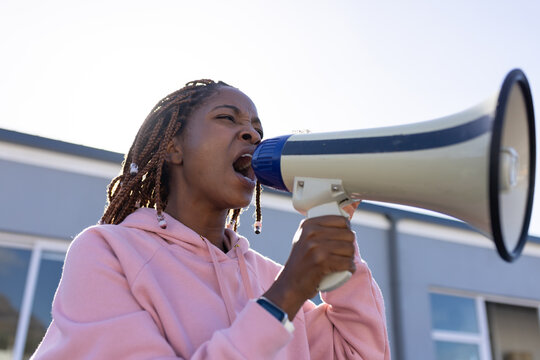 African woman shouting into beige megaphone, pink hoodie beaded braids wristband before building