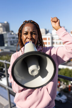 Woman wearing pink hoodie shouting into megaphone and raising clenched fist on rooftop railing