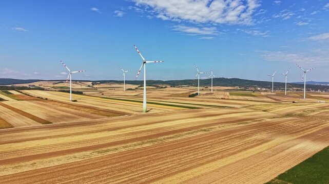 Groups of wind turbines spin slowly in a large agricultural field. The scene shows rows of crops and land extending to the horizon with a blue sky above.