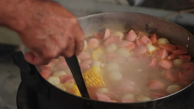 Close-up of senior cook using a ladle to stir and scoop corn, fish balls, and sausages in boiling hotpot