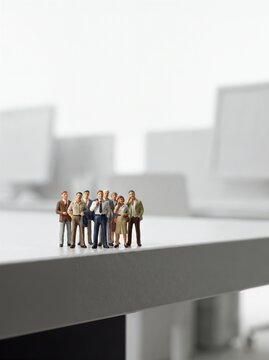 Multiple miniature business people figures standing together on a white desk edge in a bright office environment, corporate teamwork, human resources and business organization