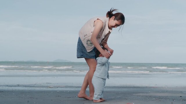  Asian mother helping her baby boy take first steps on sandy beach during family vacation.