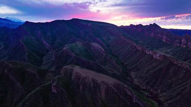 Aerial view of rugged mountain ridges and rocky valleys beneath pink sunset clouds, with dark green slopes and distant snow peaks.